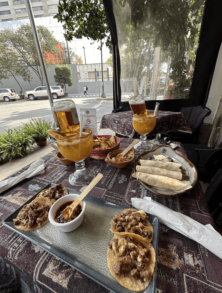 A table set outdoors with various Mexican dishes—tacos, rolled tortillas, salsas, and two large glasses of beer with upside-down bottles—rests on a decorative tablecloth beside a city street. It’s How I Travel at unique travel destinations.